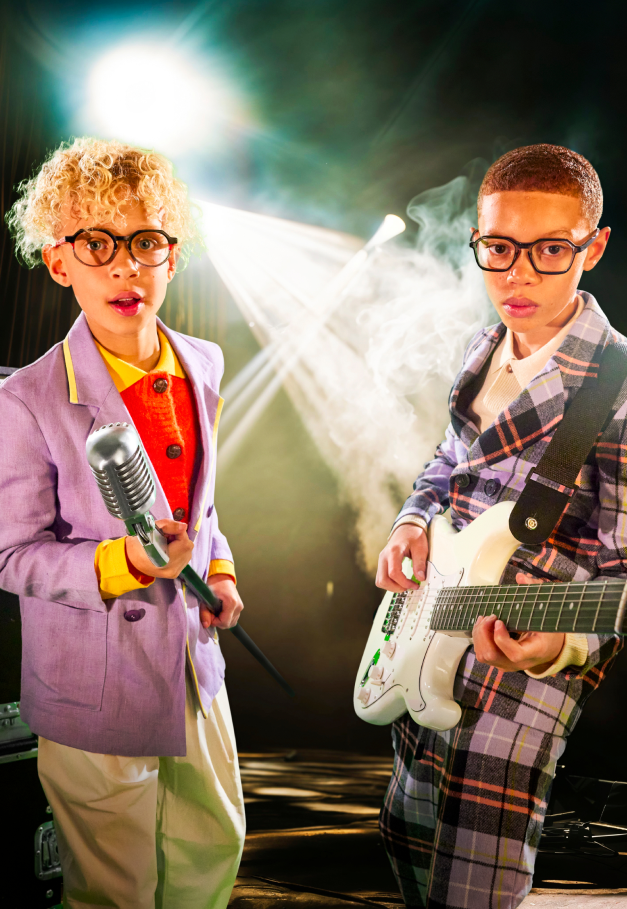 2 garçons habillés en costume coloré jouent de la musique dans un décor de concert. L'un a un micro dans les mains et l'autre une guitare électrique. Ils portent tous les 2 des lunettes de la marque de créateur Not for kids® ou NFK®.